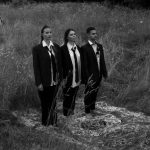 Three adults in suits stand on a patterned carpet in a grassy meadow with cows.